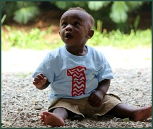 Black baby boy sitting outside, wearing a shirt reading 1