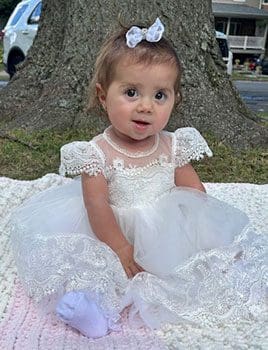 Baby Remi dressed in her baptism gown sitting under a tree